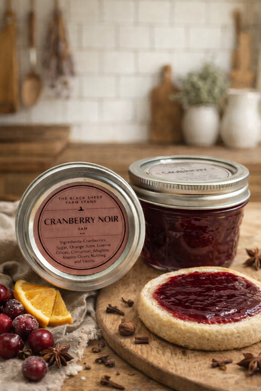 Cranberry Noir jam jar with a lid, surrounded by cranberries, an orange slice, and an English muffin with jam on a cutting board.