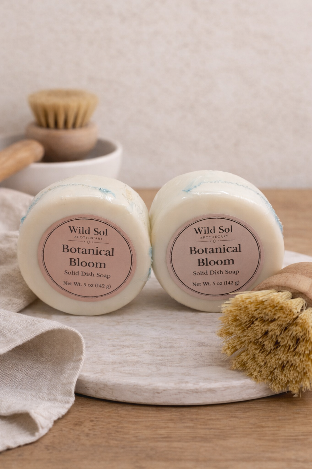 2 Solid dish soaps on a stone with a dish brush. A dish brush and towel in the background